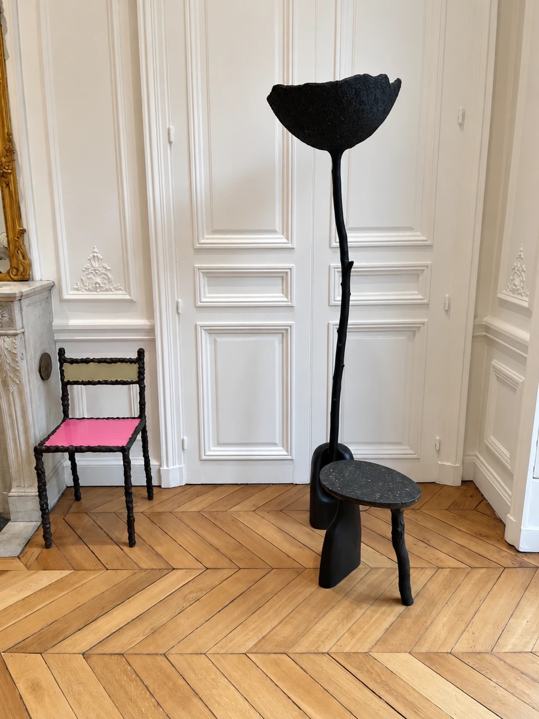 A black sculptural floor lamp with a flower-shaped shade beside a small side table and a chair with a pink seat, on a herringbone floor in front of white paneled doors