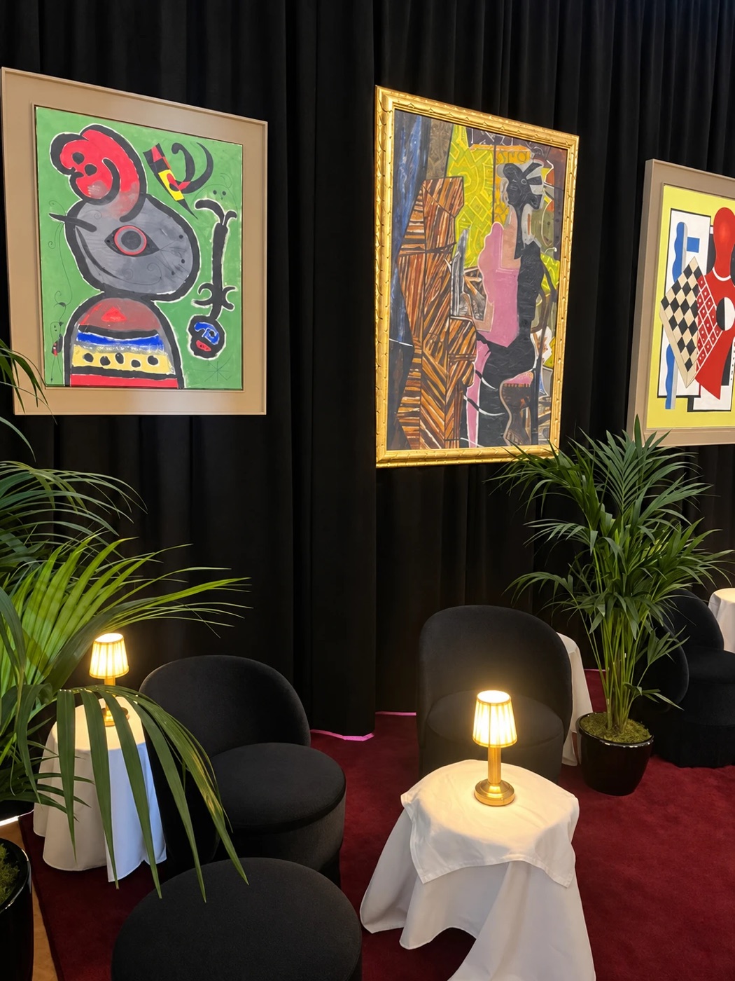 Modern paintings in gold and pale frames against dark curtains, with small glowing table lamps and potted palms