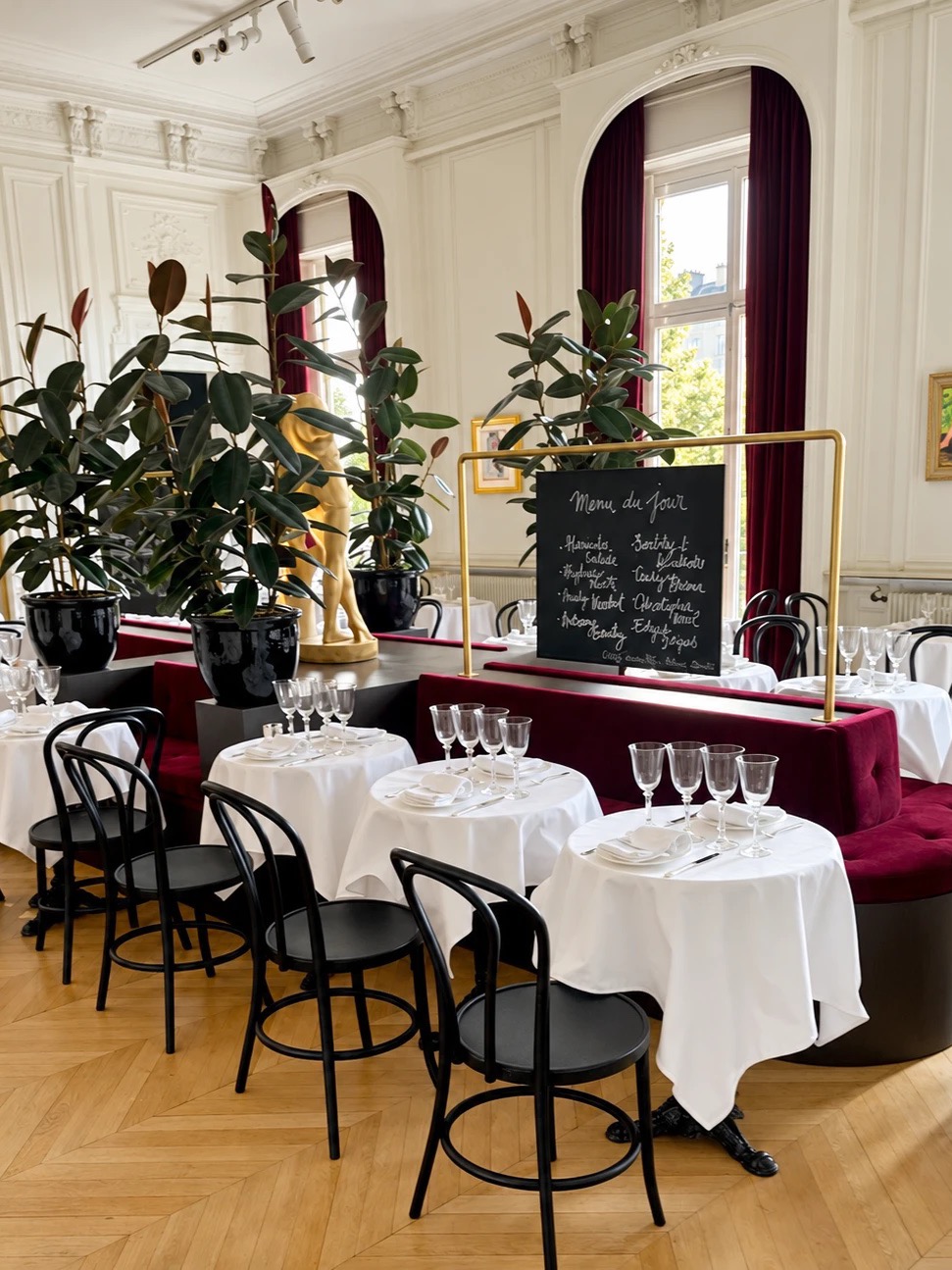 A restaurant set up in a paneled room, with round tables, crystal glasses, a burgundy banquette, and large rubber plants
