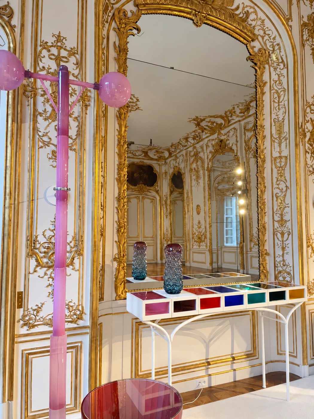 A pink glass coat stand and a colorful glass-compartment console beneath a gilded mirror in an eighteenth-century paneled room