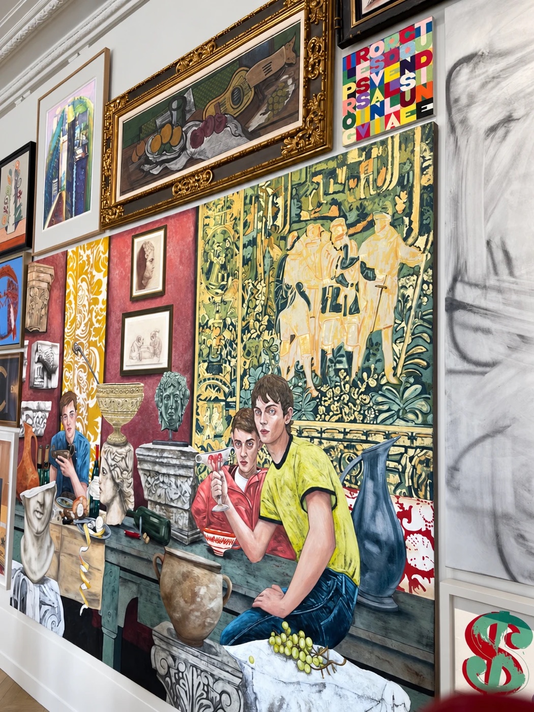 A large figurative painting of young men at a table with classical sculptures, surrounded by smaller framed works hung salon-style