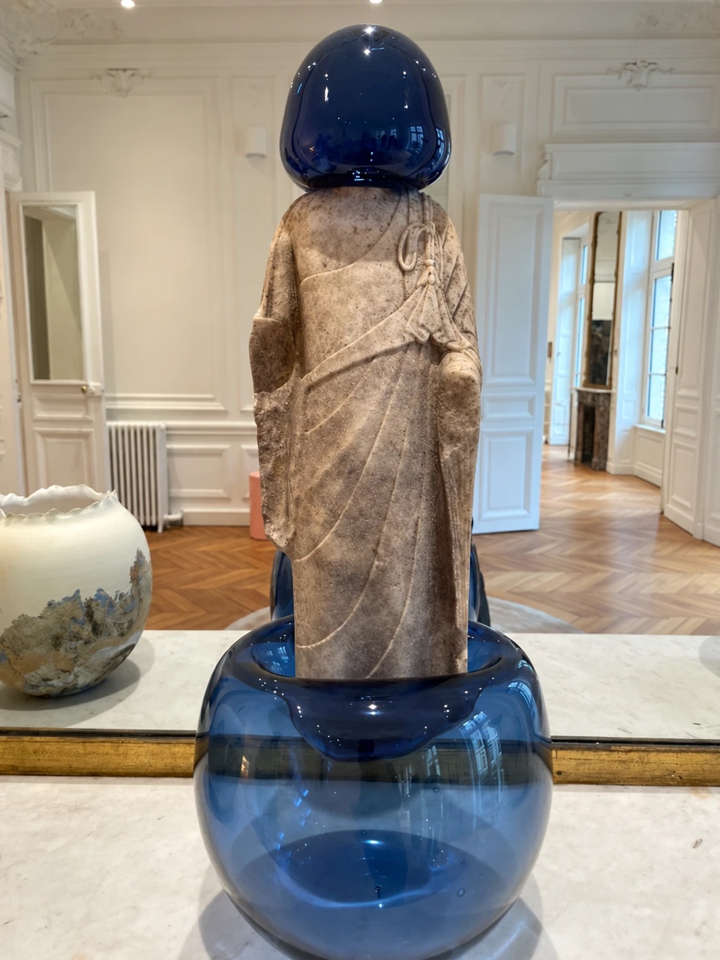 A carved stone figure with a blue glass head standing in a blue glass bowl on a marble console, beside a ceramic vase, in a paneled room