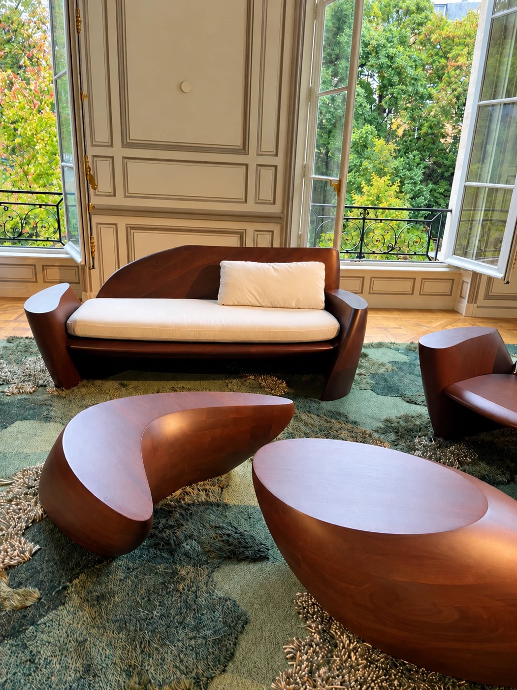 Sculptural wooden sofa and low tables on a green rug in front of open windows overlooking trees